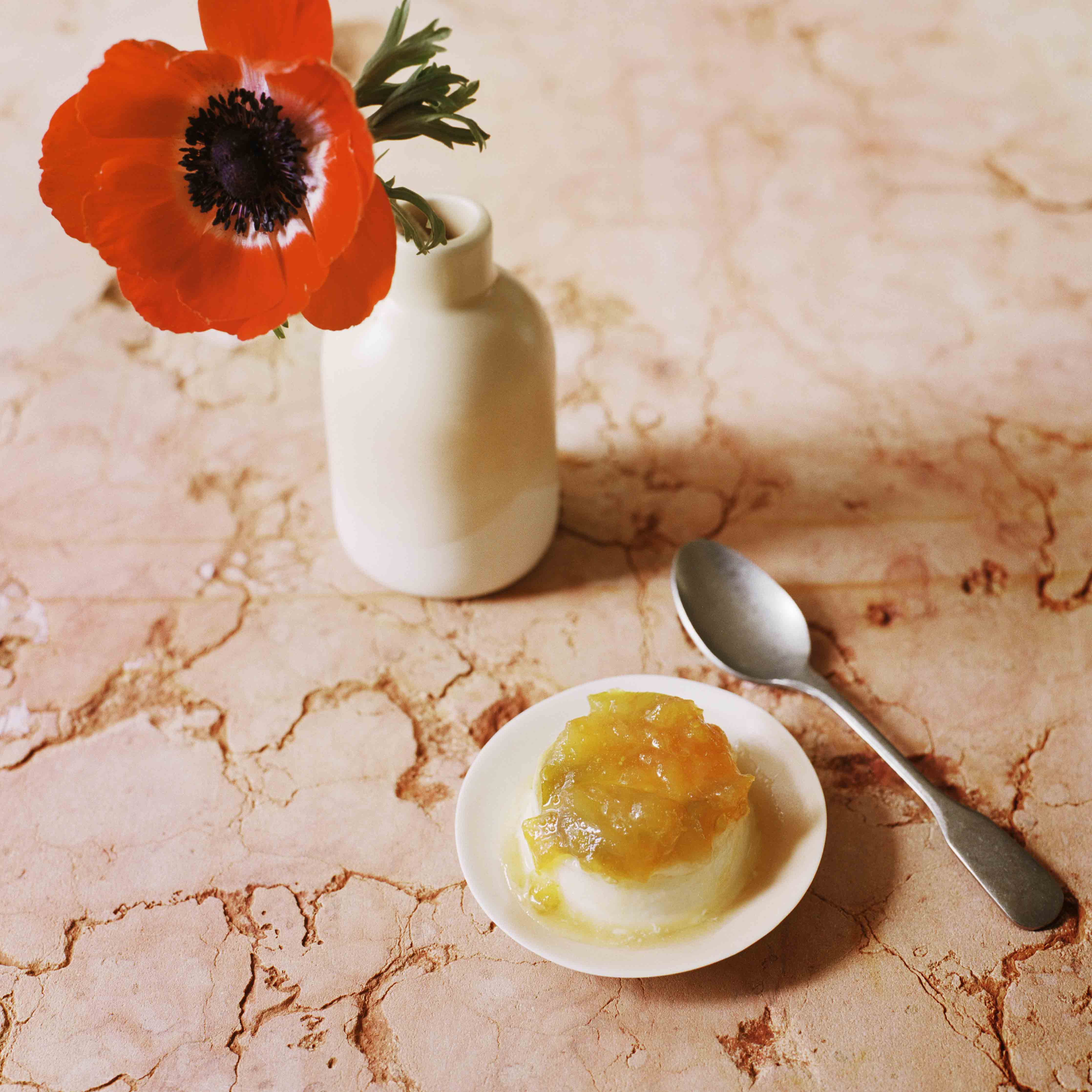 Panna cotta mit grünen Tomaten auf einem Keramikteller neben einer weissen Porzellanvase in der eine rote Mohnblume steckt