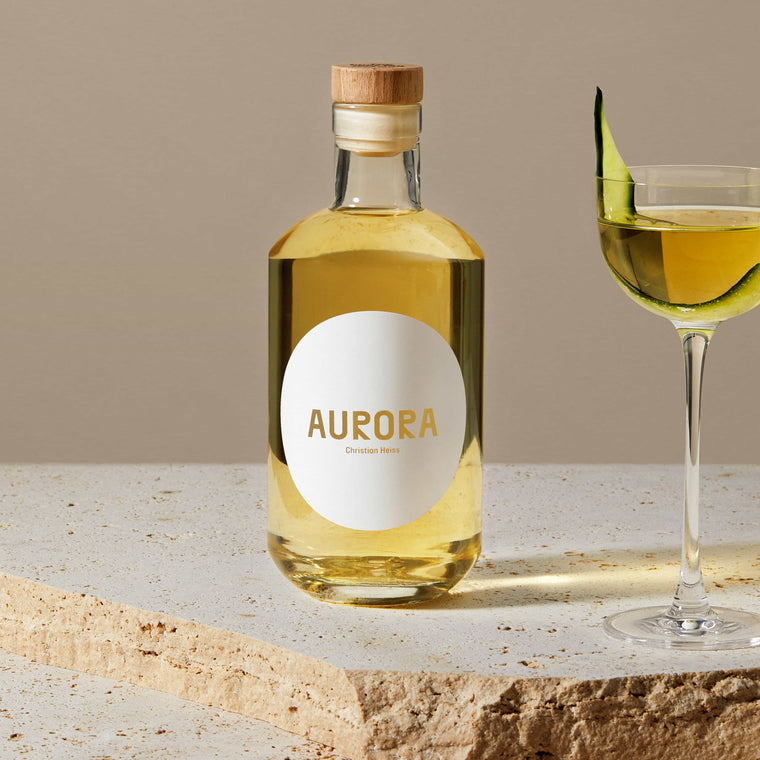Stimmungsbild der Aurora Flasche auf Travertin neben einem Cocktailglas mit Aurora und Gurkenscheibe