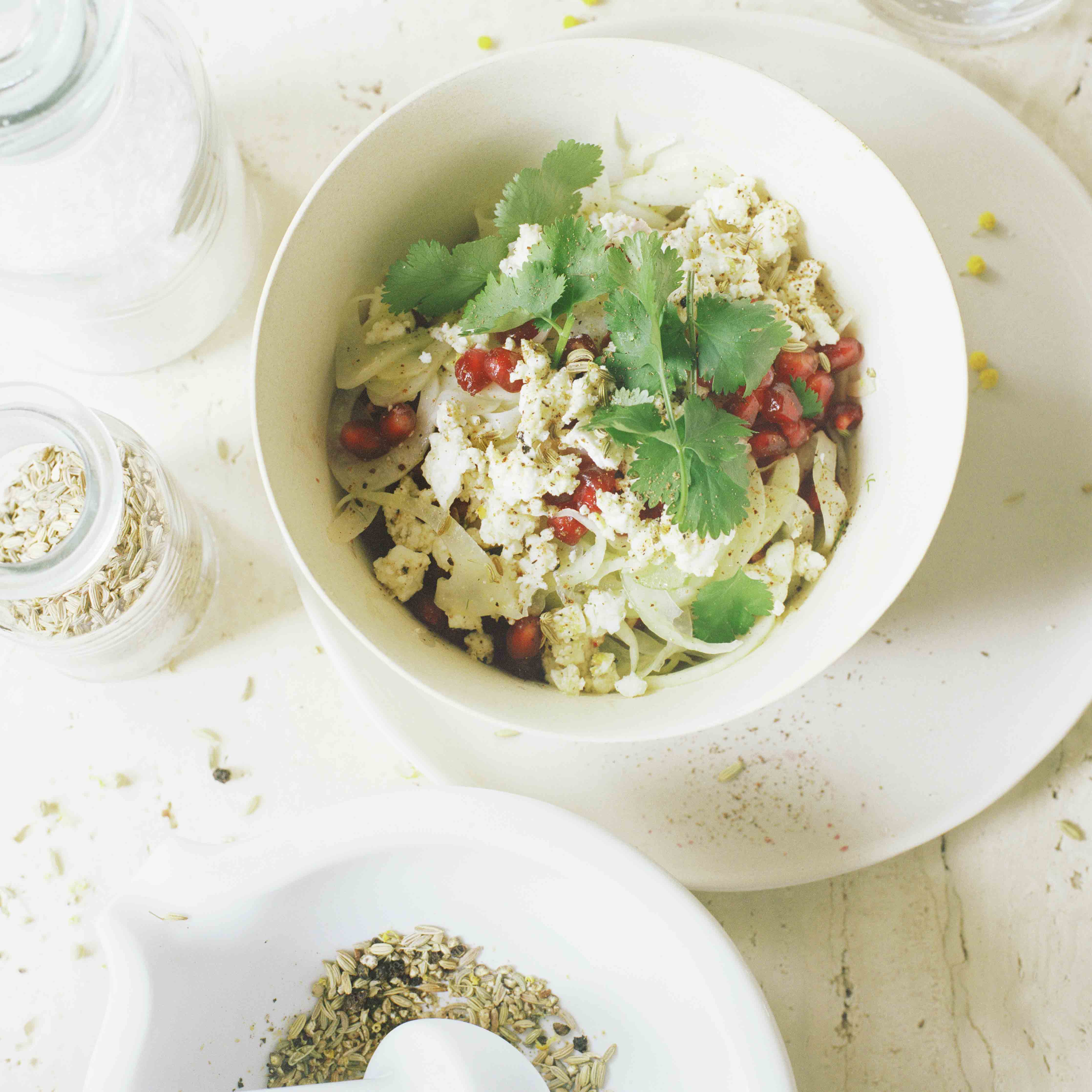 Eine weisse Schale voll Fenchelsalat mit Granatapfel und mariniertem Feta neben einem weissen Mörser aus Laborporzellan