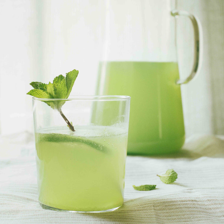 Ein klares Trinkglas von LSA gefüllt mit grüner Gurken-Pfefferminz-Limonade und einem Glaskrug von Trendglas Jena im Hintergrund