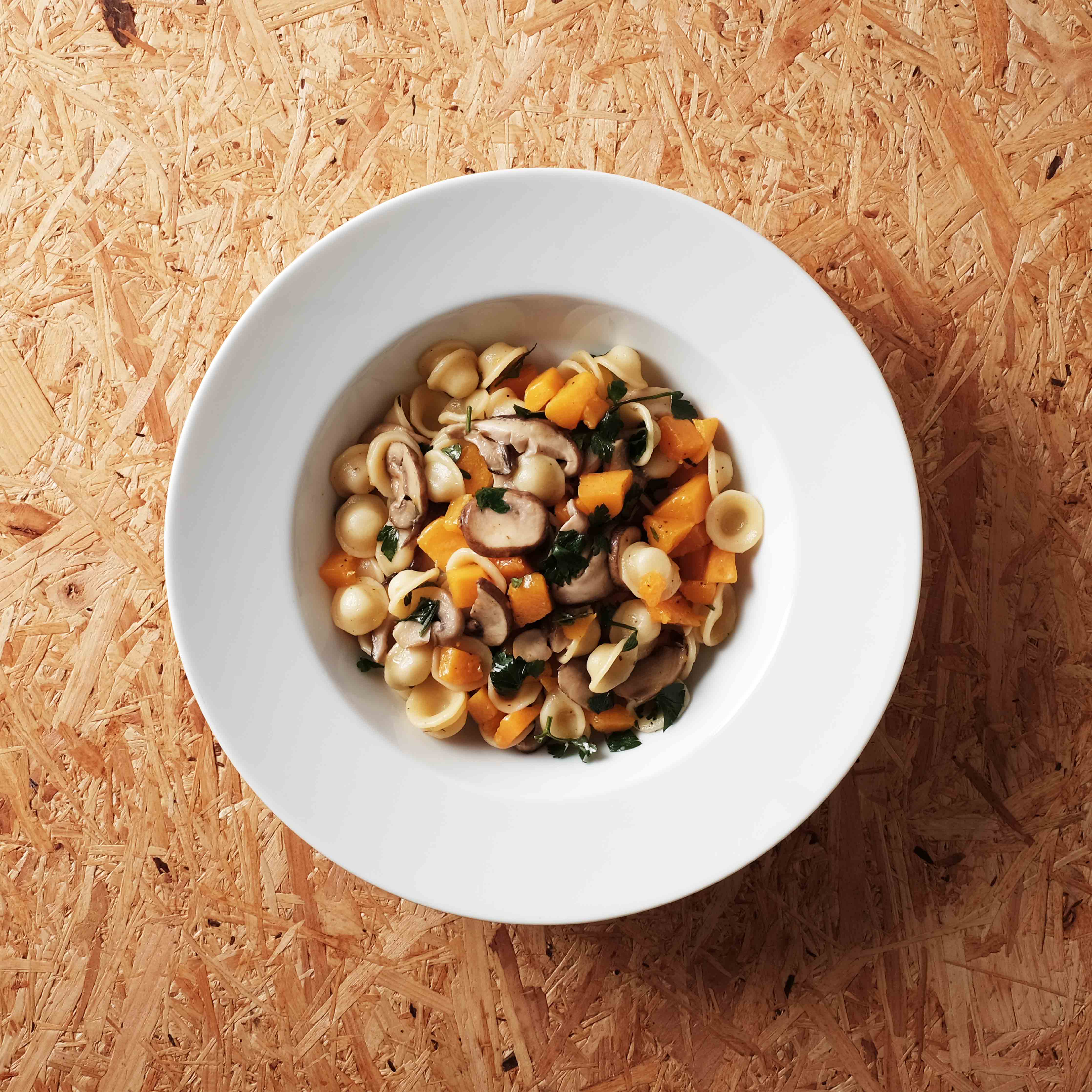 Weisser Teller mit Orecchiette, Kürbisstücken, Pilzen und Petersilie auf einer Holzplatte