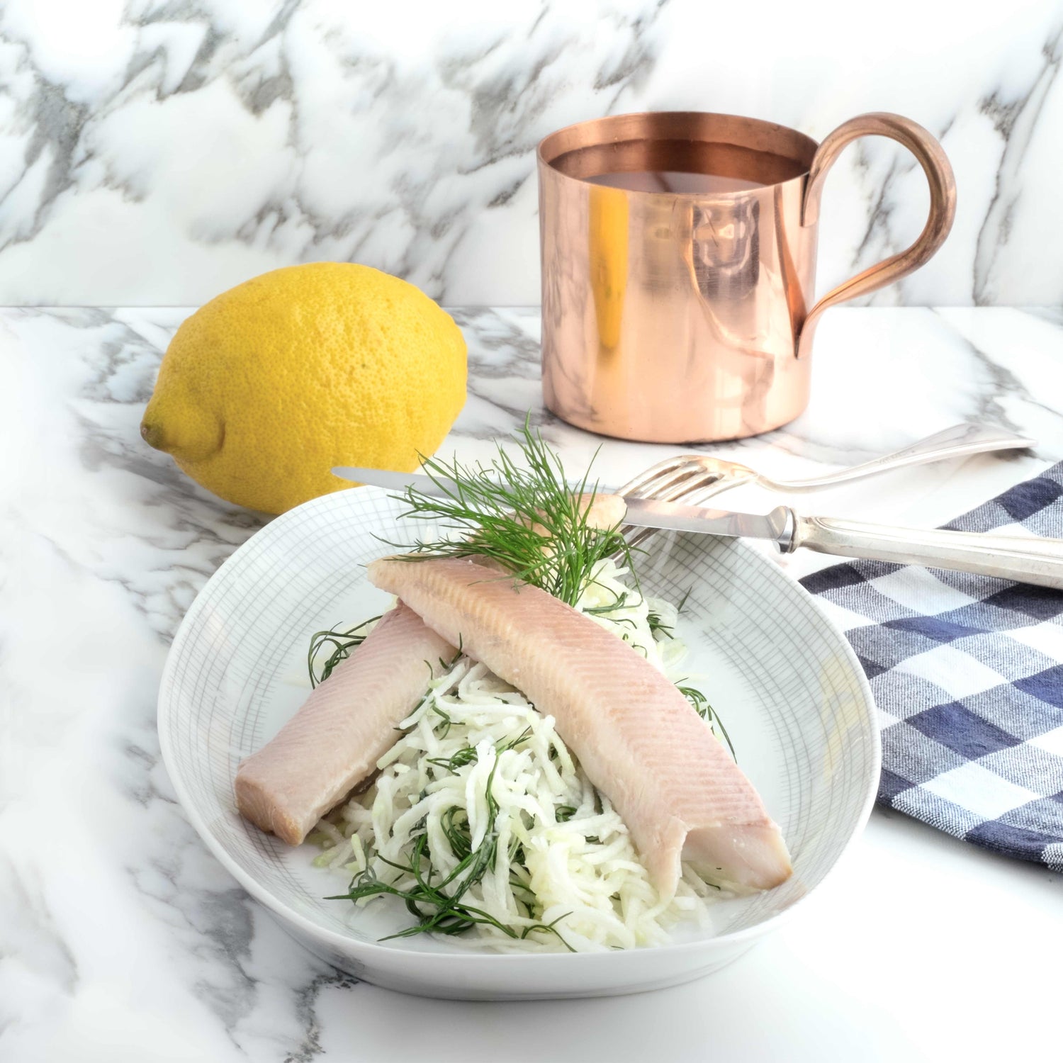 Platte mit Forelle, Kohlrabisalat und Dill-Garnitur, dahinter ein Kupferbecher stehend und eine ganze Zitrone