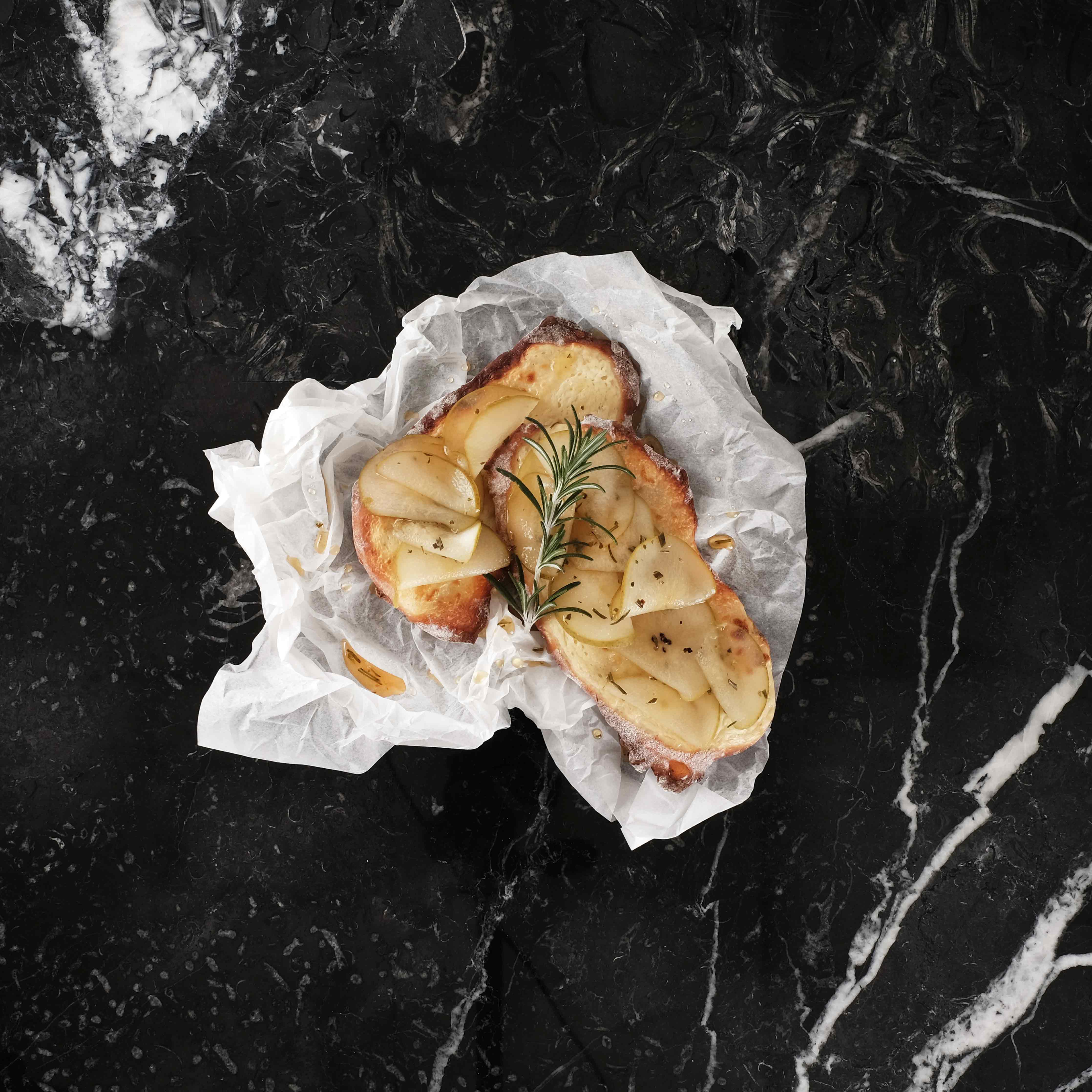 Zwei Stück Focaccia mit Birne belegt und einem Zweig Thymian garniert, präsentiert auf schwarzem Marmor