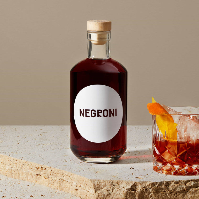 Stimmungsbild der Negroni Flasche auf Travertin neben einem Tumbler mit Negroni, Eis und Orange