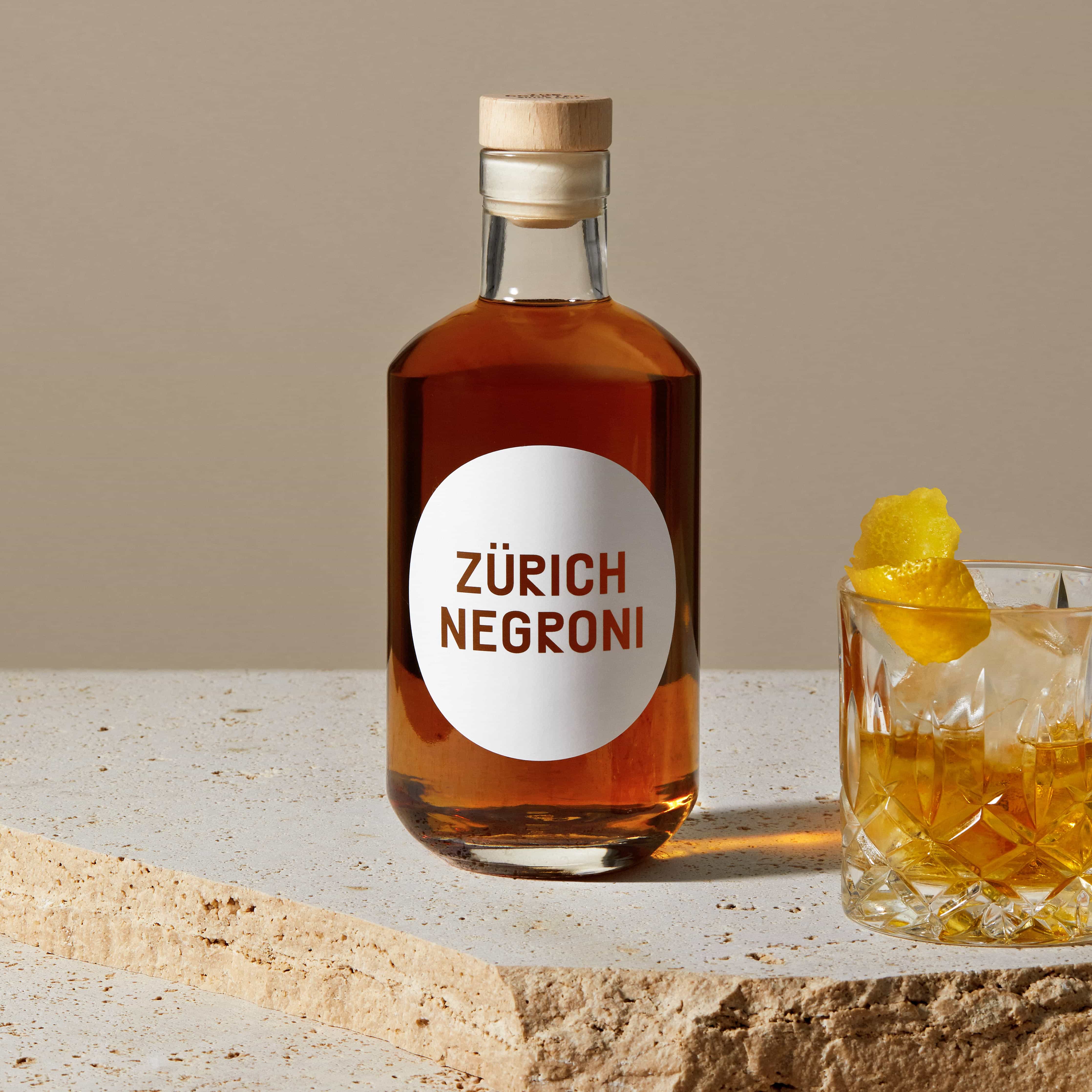 mood: Stimmungsbild der Zürich Negroni Flasche auf Travertin neben einem Tumbler mit Negroni, Eis und Orange